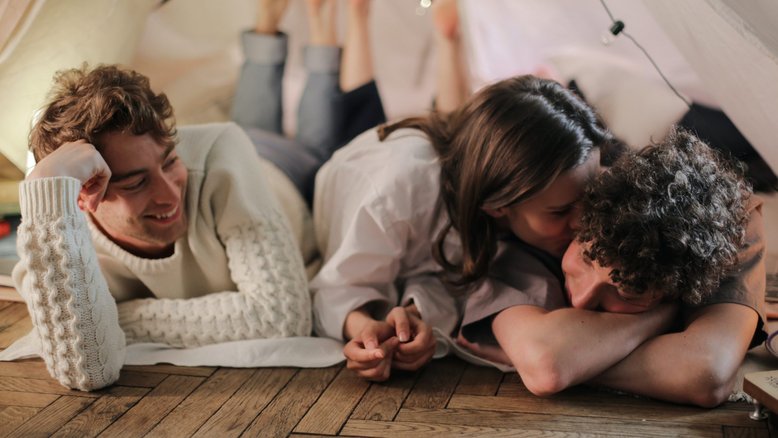 relacionamento aberto, dois homens e uma mulher deitados de bruços. a mulher está beijando um dos homens enquanto o outro sorri