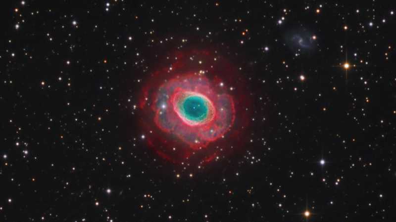 A Nebulosa do Anel (M57) está a cerca de 2.000 anos-luz de distância na Constelação de Lira. O anel central tem cerca de um ano-luz de diâmetro, mas esta exposição notavelmente profunda explora os filamentos em loop de gás brilhante que se estendem muito mais longe da estrela central da nebulosa. O material brilhante não vem de planetas. Em vez disso, a mortalha gasosa representa as camadas externas expelidas de uma estrela moribunda parecida com o Sol. A cor turquesa é decorrência da emissão de átomos de oxigênio em temperaturas mais altas dentro do anel