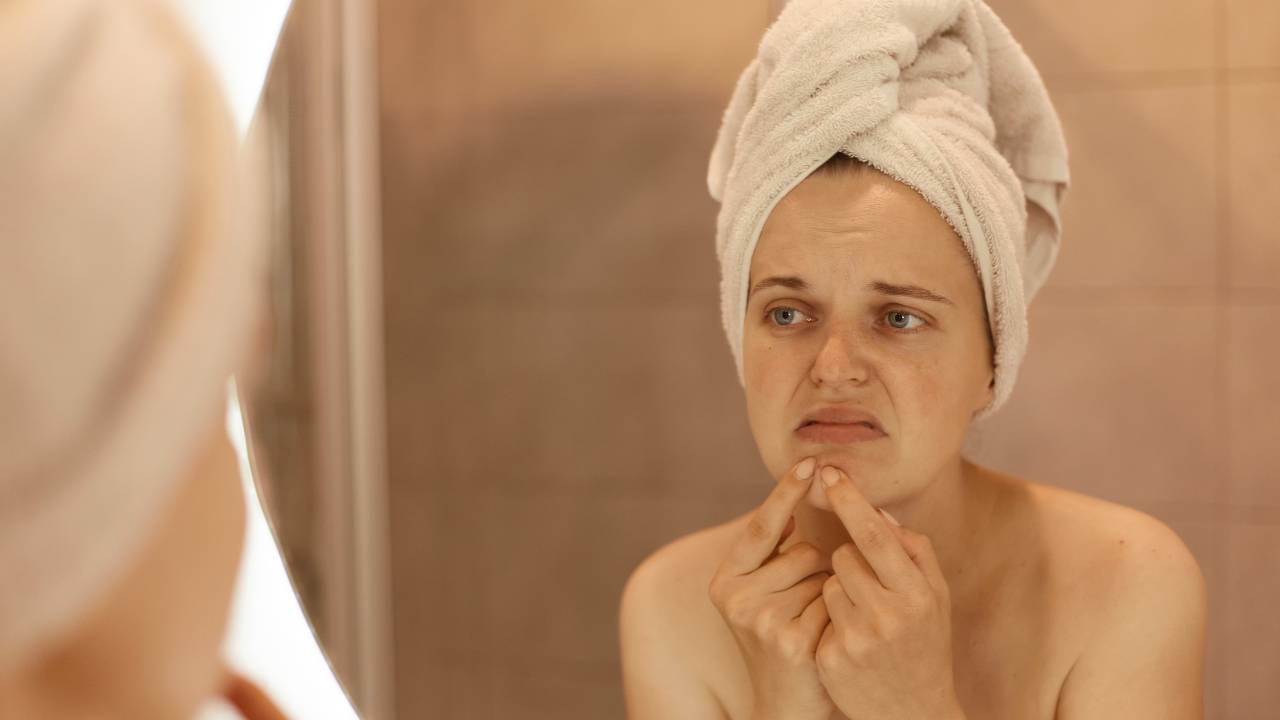 mulher no espelho cutucando acne, como a ansiedade prejudica a pele