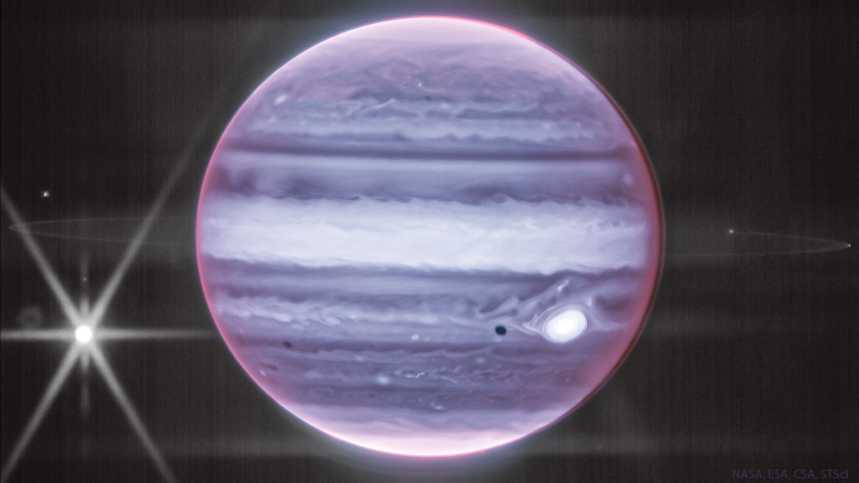 Esta imagem mostra Júpiter, em luz infravermelha, registrado pelo Telescópio Espacial James Webb. Junto ao gigante gasoso e suas nuvens, vemos também seu anel. A famosa Grande Mancha Vermelha do planeta aparece em cor clara, à direita. A grande lua de Júpiter, Europa, está à esquerda, no centro dos picos de difração (as formas brilhantes e pontiagudas), à esquerda. Já a sombra de Europa pode ser vista ao lado da famosa mancha de Júpiter. O pequeno ponto próximo ao anel, à esquerda, é Tebe, outra lua que orbita o gigante gasoso, assim como Métis, à direita