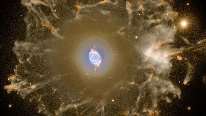 Localizada a cerca de 3,3 mil anos-luz, na direção da Constelação de Draco, a Nebulosa Olho de Gato (NGC 6543) é uma das mais conhecidas nebulosas planetárias. Suas simetrias são notadas na região central desta imagem com cores falsas, que foi processada especialmente para revelar o halo de material gasoso que a circunda, com mais de três anos-luz de diâmetro. Feita com dados do Telescópio Óptico Nórdico, nas Ilhas Canárias, a imagem composta mostra a emissão estendida da nebulosa. Embora considere-se que a fase de nebulosa planetária dure cerca de 10 mil anos, estima-se que as porções filamentosas externas desse halo tenham entre 50 mil e 90 mil anos. As nebulosas planetárias representam a fase final da vida de uma estrela parecida com o Sol