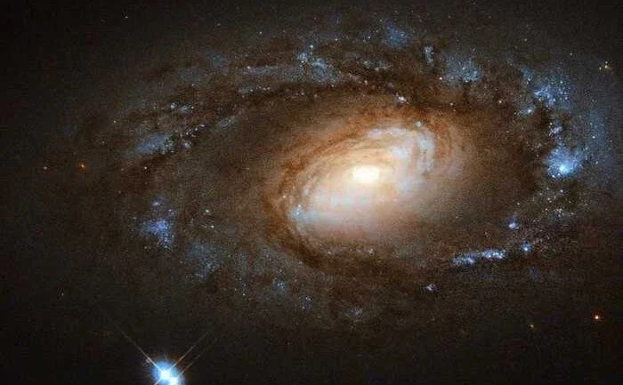 As galáxias, assim como os humanos, não são todas supermodelos. Esta pequena espiral, conhecida como NGC 4102, tem um tipo diferente de beleza, com seus braços espirais bem enrolados e aparência discreta, mas charmosa. NGC 4102 fica na Constelação da Ursa Maior, a aproximadamente 60 milhões de anos-luz da Terra. Ela contém uma região de explosão estelar, onde as estrelas estão sendo criadas a uma taxa muito diferente da média. Isso acontece dentro de um pequeno disco giratório, com cerca de mil anos-luz de diâmetro e uma massa cerca de três bilhões de vezes a massa do Sol