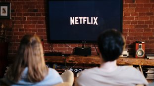 Mulher e homem são vistos de costas, olhando para uma TV com o símbolo da Netflix