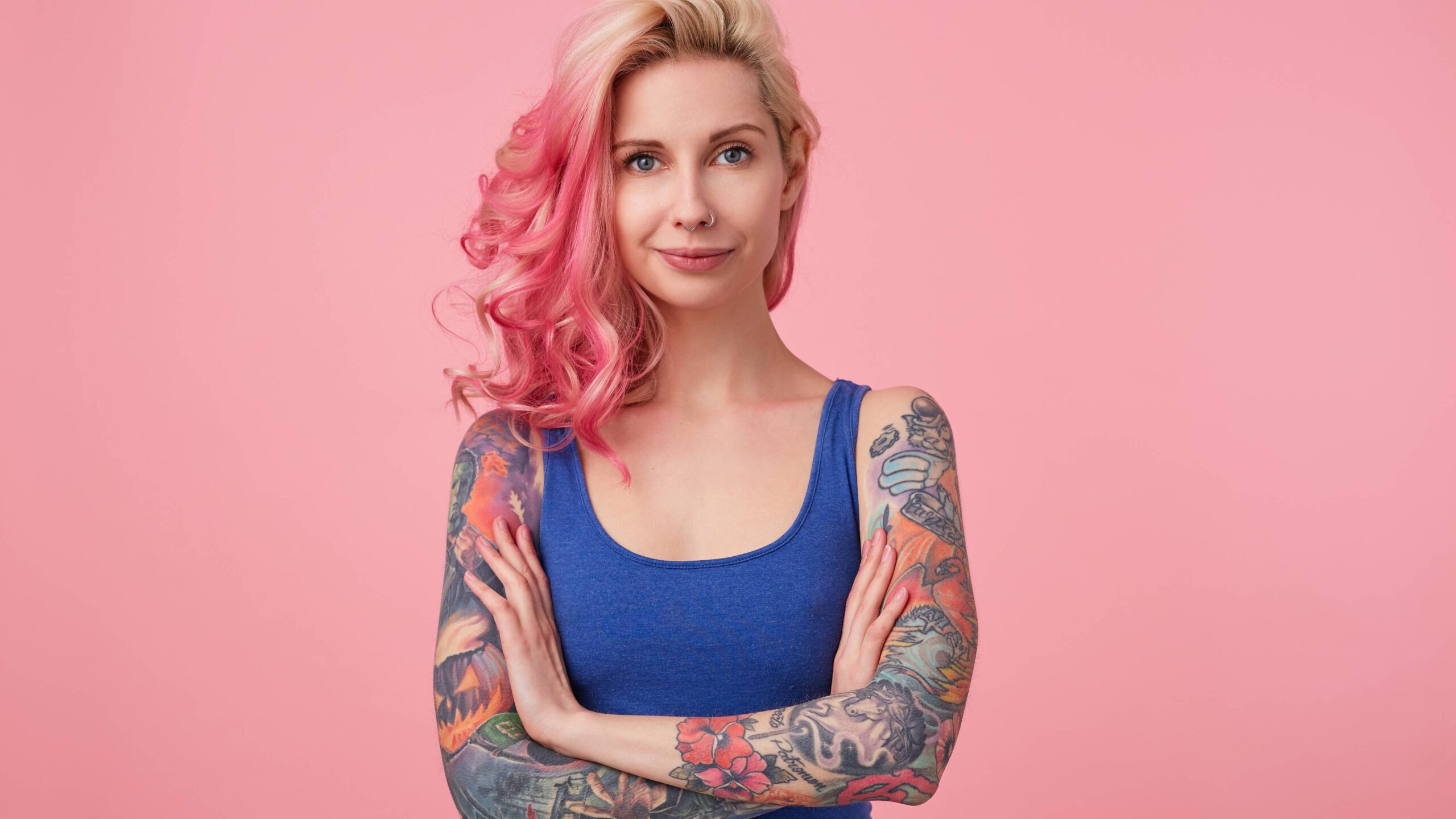 Mulher jovem com braços tatuados cruzados, regata azul e cabelo loiro com mechas rosas em fundo rosa