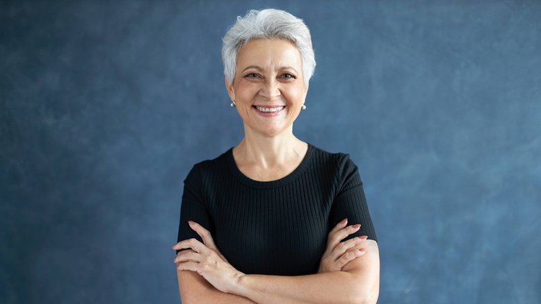 mulher de cabelos brancos e roupa preta cruzando os braços e sorrindo para a câmera