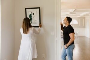 Olhar fotos de sua pessoa amada pode aumentar seu amor; entenda 2 mulher pendurando imagem de casamento na parede, marido olhando para ela, amor