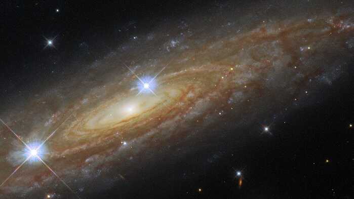 Nesta imagem, vê-se a galáxia espiral UGC 11537, com suas faixas brilhantes de estrelas e também as nuvens escuras de poeira que se espalham por toda a galáxia. UGC 11537 está a 230 milhões de anos-luz de distância, na Constelação da Águia, e fica perto do plano da Via Láctea, fazendo com que estrelas em primeiro plano de nossa própria galáxia se infiltrassem na imagem, como as duas proeminentes que aparecem logo à frente. Essas estrelas brilhantes em primeiro plano são cercadas por picos de difração, causados pela interação da luz com a estrutura interna do Telescópio Espacial Hubble, responsável por este registro