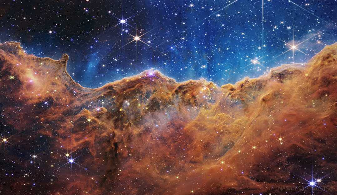Os ventos destrutivos e a luz energética das estrelas no aglomerado aberto NGC 3324 erodiram parte de uma montanha de poeira interestelar escura, na parte norte da Nebulosa de Carina. Várias dessas estrelas estão visíveis no topo desta imagem detalhada, feita pelo tecnológico Telescópio Espacial James Webb. As falésias irregulares em destaque estão na parte de Carina conhecida como Nebulosa Gabriela Mistral, que recebeu esse nome em homenagem à famosa poeta chilena de mesmo nome, pois essas formações lembram o seu perfil facial, dependendo do ângulo de visão. Esses penhascos nebulares estão a cerca de 7,6 mil anos-luz de distância, na direção da austral Constelação de Carina