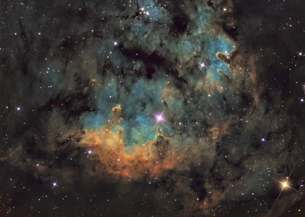 Estrelas quentes e jovens em conjunto a pilares cósmicos de gás e poeira parecem se aglomerar em NGC 7822. Na borda de uma nuvem molecular gigante na Constelação de Cepheus, essa região de formação estelar brilhante fica a cerca de 3 mil anos-luz de distância. Nesta colorida paisagem telescópica, vê-se a emissão de oxigênio atômico, hidrogênio e enxofre em tons de azul, verde e vermelho. A emissão atômica é alimentada pela radiação energética das estrelas quentes centrais. As estrelas ainda podem estar se formando dentro dos pilares por colapso gravitacional