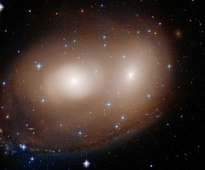 O que parecem ser dois olhos brilhantes e um sorriso torto nesta imagem, na verdade, são os estágios iniciais de uma colisão entre duas galáxias. Ambas estão na Constelação do Cão Maior, a cerca de 120 milhões de anos-luz de distância. A cor geral, que varia nos tons de abóbora, corresponde ao brilho das estrelas vermelhas envelhecidas nas duas galáxias, catalogadas como NGC 2292 e NGC 2293. O braço que parece formar um “sorriso” pode ser apenas o começo do processo de reconstrução de uma galáxia espiral, apontam as pesquisas, e provavelmente se formou quando o gás interestelar foi comprimido