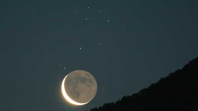 A Lua Minguante crescente compartilha o céu oriental sobre Menton, na França, com as estrelas irmãs do aglomerado das Plêiades, na Constelação de Touro. A imagem foi registrada em uma manhã de junho de 2006. Imagens astronômicas das Plêiades geralmente mostram as nebulosas de reflexão azul do aglomerado, mas elas estão aqui ofuscadas pelo luar. Enquanto a Lua Minguante está superexposta, as características de sua superfície podem ser vistas por meio da luz cinérea. Ela é resultado do brilho refletido pela Terra, iluminada pelo Sol, que revela a porção obscurecida de nosso satélite natural