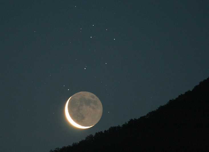 A Lua Minguante crescente compartilha o céu oriental sobre Menton, na França, com as estrelas irmãs do aglomerado das Plêiades, na Constelação de Touro. A imagem foi registrada em uma manhã de junho de 2006. Imagens astronômicas das Plêiades geralmente mostram as nebulosas de reflexão azul do aglomerado, mas elas estão aqui ofuscadas pelo luar. Enquanto a Lua Minguante está superexposta, as características de sua superfície podem ser vistas por meio da luz cinérea. Ela é resultado do brilho refletido pela Terra, iluminada pelo Sol, que revela a porção obscurecida de nosso satélite natural