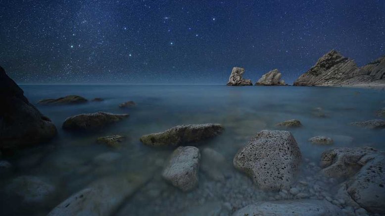 A Galáxia de Andrômeda, a 2,5 milhões de anos-luz de distância, é o objeto mais distante visível a olho a partir da Terra. Outros objetos aparentes no céu noturno, como estrelas, aglomerados e nebulosas, estão entre centenas e milhares de anos-luz de distância, localizados dentro da nossa Via Láctea. Também conhecida como M31, a Galáxia de Andrômeda é a mancha tênue, perto do centro superior desta paisagem celeste, captada na região de Monte Conero, na costa do Mar Adriático, na Itália. Como as estrelas se arrastavam acima do horizonte enquanto a foto era feita, exposições separadas foram combinadas para retratar esta cena sublime