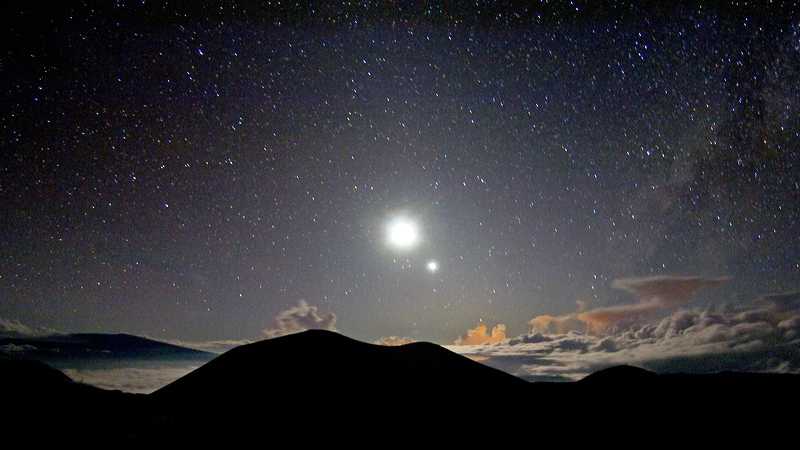 Do alto do pico Mauna Kea, na Ilha Grande do Havaí, é possível que se tenha uma visão noturna espetacular. A uma altitude de mais de 4 mil metros, o topo da montanha é recortado nesta paisagem registrada perto do anoitecer, em dezembro de 2005. O pico vulcânico se eleva acima das nuvens de tempestade, iluminadas pela Lua brilhante. O planeta Vênus está se pondo perto da Lua, em sua fase como estrela vespertina. A cena também inclui uma leve faixa leitosa, que se estende do horizonte para o céu ao longo da borda direita do quadro, retratando o disco de estrelas da nossa galáxia, a Via Láctea, junto a nuvens de poeira cósmica