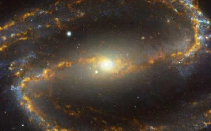 A galáxia NGC 1300 está localizada a aproximadamente 61 milhões de anos-luz da Terra, na Constelação de Eridanus. Ela é uma galáxia espiral, com uma barra de estrelas e gás –– visto aqui cruzando a imagem horizontalmente –– e um anel central de intensa formação estelar. Esta imagem é uma combinação de observações realizadas em diferentes cores –– ou comprimentos de onda –– de luz. O tom caramelo e com brilho dourado corresponde a nuvens de gás molecular, a matéria-prima a partir da qual as estrelas se formam. As regiões azuladas, ao fundo, revelam a distribuição de estrelas mais velhas e já formadas
