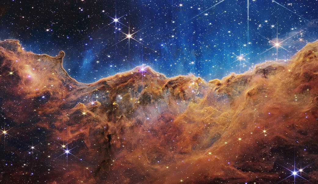 Esta paisagem semelhante a “montanhas” salpicadas de estrelas brilhantes é, na verdade, a borda de uma jovem região de formação de estrelas, chamada NGC 3324, a cerca de 7,6 mil anos-luz de distância. Ela representa o canto noroeste da famosa Nebulosa Carina (NGC 3372), localizada na austral Constelação de Carina (“A Quilha”), que também abriga a estrela supergigante, ativa e instável, Eta Carinae. Neste registro feito em luz infravermelha, com o Telescópio Espacial James Webb, revelou-se, pela primeira vez, áreas anteriormente invisíveis. Estes “picos” mais altos têm cerca de 7 anos-luz de altura, sendo que a área cavernosa foi esculpida pela intensa radiação ultravioleta e ventos estelares de estrelas jovens extremamente massivas e quentes