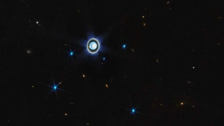 Esta visão ampla, registrada pelo Telescópio Espacial James Webb, mostra o planeta Urano rodeado por seis de suas 27 luas conhecidas. Vistas aqui como pequenos pontos azuis e brilhantes, estão Ariel, Puck, Miranda, Umbriel, Titânia e Oberon. Os seus demais satélites naturais são muito pequenos e fracos para que fossem notados nesta exposição de curta duração. Os pontos mais amarelados da imagem representam variados objetos de fundo, incluindo galáxias vizinhas à Via Láctea