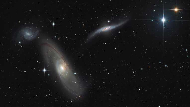 Nesta imagem, vê-se um trio de galáxias em interação (Arp 286), a quase 90 milhões de anos-luz de distância, na direção da Constelação de Virgem. À direita, há também duas estrelas pontiagudas da nossa Via Láctea, em primeiro plano. Com braços espirais arrebatadores e faixas de poeira obscuras, NGC 5566 é enorme, com cerca de 150 mil anos-luz de diâmetro. Logo acima dela, está a pequena e azulada NGC 5569. Perto do centro, a terceira galáxia, NGC 5560, é multicolorida e está aparentemente distorcida, em função da interação gravitacional com NGC 5566. Tais interações cósmicas são agora entendidas como uma parte da evolução das galáxias no Universo