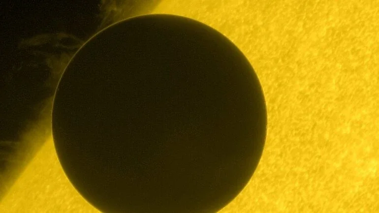 Nesta imagem, registrada a partir da espaçonave Hinode, da NASA, em junho de 2012, o planeta Vênus, em trânsito, cruza a borda do Sol. O tempo de cruzamento durante esses raros trânsitos é utilizado historicamente para triangular a distância de Vênus e, com isso, determinar o valor da distância entre a Terra e o Sol, chamada de unidade astronômica. Em meio a esse procedimento de medição, pode-se notar também a turbulenta superfície solar, com proeminências elevadas por campos magnéticos distorcidos. O fino anel de luz, em torno da silhueta escura de Vênus, representa a luz solar refratada por sua espessa atmosfera