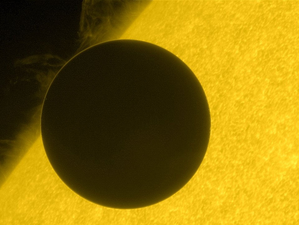 Nesta imagem, registrada a partir da espaçonave Hinode, da NASA, em junho de 2012, o planeta Vênus, em trânsito, cruza a borda do Sol. O tempo de cruzamento durante esses raros trânsitos é utilizado historicamente para triangular a distância de Vênus e, com isso, determinar o valor da distância entre a Terra e o Sol, chamada de unidade astronômica. Em meio a esse procedimento de medição, pode-se notar também a turbulenta superfície solar, com proeminências elevadas por campos magnéticos distorcidos. O fino anel de luz, em torno da silhueta escura de Vênus, representa a luz solar refratada por sua espessa atmosfera