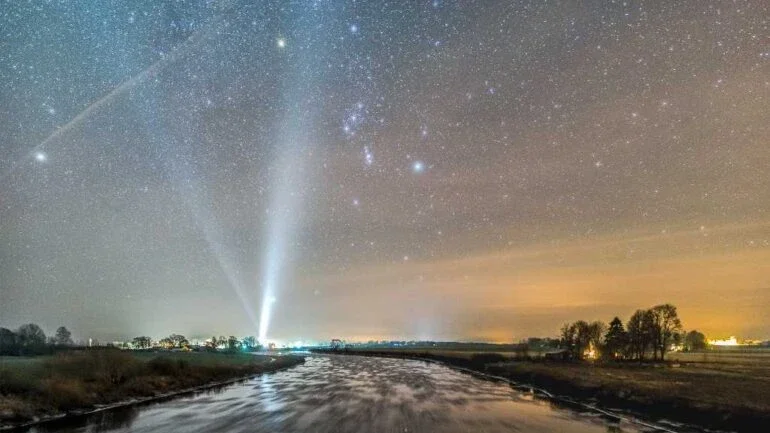 O gelo se acumula ao longo do rio Lielupe, localizado perto da vila de Stalgene, na Letônia, nesta paisagem noturna do inverno boreal. Mesmo em movimento, a água gelada ainda reflete um céu estrelado. O panorama bem planejado, centrado na Constelação de Orion, mira a direção Sul. Formações congeladas deixam rastros na imagem, enquanto refletem as estrelas. As luzes da vila, ao longo do horizonte, incluem feixes de luz da igreja da comunidade local