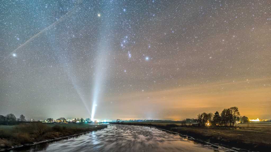 O gelo se acumula ao longo do rio Lielupe, localizado perto da vila de Stalgene, na Letônia, nesta paisagem noturna do inverno boreal. Mesmo em movimento, a água gelada ainda reflete um céu estrelado. O panorama bem planejado, centrado na Constelação de Orion, mira a direção Sul. Formações congeladas deixam rastros na imagem, enquanto refletem as estrelas. As luzes da vila, ao longo do horizonte, incluem feixes de luz da igreja da comunidade local