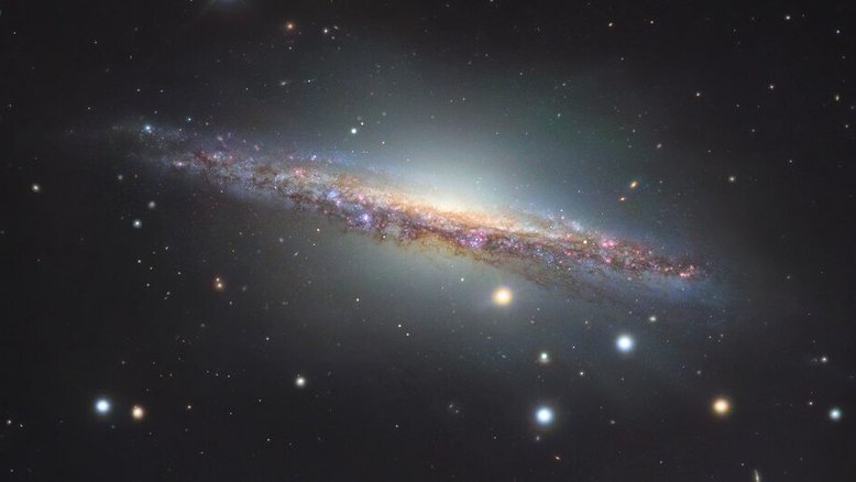 A galáxia espiral NGC 1055 é o membro dominante de um pequeno grupo de galáxias a 60 milhões de anos-luz de distância, na direção da Constelação de Cetus. Vista de lado, ela se estende por mais de 100 mil anos-luz, sendo um pouco maior que a nossa Via Láctea. As estrelas coloridas da imagem estão em primeiro plano, dentro da nossa galáxia. As regiões rosadas ao longo do disco fino de NGC 1055 indicam a formação de estrelas. Este registro também revela um halo que se estende acima e abaixo de seu bulbo central. Atado a estruturas tênues, ele indica provavelmente a mistura de detritos de uma galáxia satélite destruída pela espiral maior, há cerca de 10 bilhões de anos