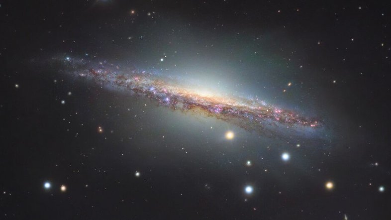 A galáxia espiral NGC 1055 é o membro dominante de um pequeno grupo de galáxias a 60 milhões de anos-luz de distância, na direção da Constelação de Cetus. Vista de lado, ela se estende por mais de 100 mil anos-luz, sendo um pouco maior que a nossa Via Láctea. As estrelas coloridas da imagem estão em primeiro plano, dentro da nossa galáxia. As regiões rosadas ao longo do disco fino de NGC 1055 indicam a formação de estrelas. Este registro também revela um halo que se estende acima e abaixo de seu bulbo central. Atado a estruturas tênues, ele indica provavelmente a mistura de detritos de uma galáxia satélite destruída pela espiral maior, há cerca de 10 bilhões de anos