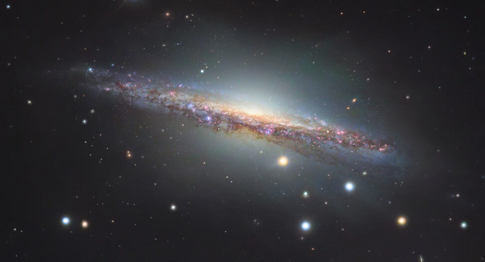 A galáxia espiral NGC 1055 é o membro dominante de um pequeno grupo de galáxias a 60 milhões de anos-luz de distância, na direção da Constelação de Cetus. Vista de lado, ela se estende por mais de 100 mil anos-luz, sendo um pouco maior que a nossa Via Láctea. As estrelas coloridas da imagem estão em primeiro plano, dentro da nossa galáxia. As regiões rosadas ao longo do disco fino de NGC 1055 indicam a formação de estrelas. Este registro também revela um halo que se estende acima e abaixo de seu bulbo central. Atado a estruturas tênues, ele indica provavelmente a mistura de detritos de uma galáxia satélite destruída pela espiral maior, há cerca de 10 bilhões de anos