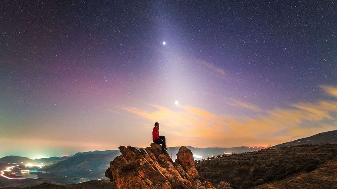 Qual é a causa da faixa de luz incomum que se estende a partir do horizonte? Poeira em órbita do Sol. Há momentos do ano nos quais uma faixa de poeira que reflete a luz solar aparece depois do poente ou antes do nascente, em um fenômeno chamado de luz zodiacal. Esta imagem foi feita em meados de fevereiro, no Parque Nacional de Sierra Nevada, na Espanha. Nela, vê-se a luz zodiacal junto aos brilhantes planetas Júpiter (acima) e Vênus (abaixo)