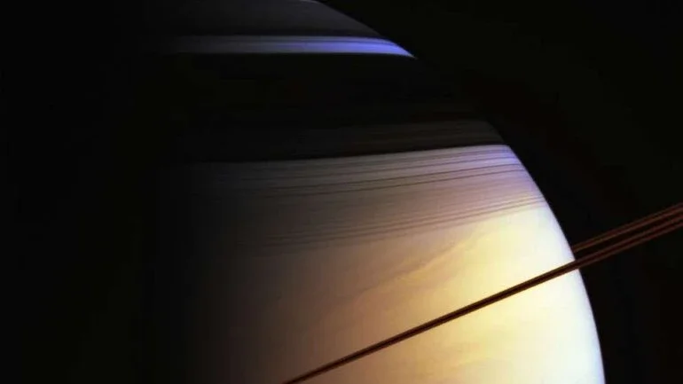 O que cria as cores de Saturno? A imagem em destaque exagera apenas um pouco o que um humano veria se estivesse perto do gigante dos anéis. Registrada em 2005, pela espaçonave Cassini, ela retrata os majestosos anéis de Saturno como uma linha curva, quase marrom, devido ao seu brilho infravermelho. Os anéis mostram melhor sua estrutura complexa nas sombras escuras, na parte superior do planeta. O Hemisfério Norte de Saturno parece azul pela mesma razão que os céus da Terra mostram-se azulados: as moléculas nas porções sem nuvens da atmosfera espalham melhor a luz azul do que a vermelha. Ao olhar profundamente nas nuvens de Saturno, no entanto, a tonalidade dourada natural torna-se dominante
