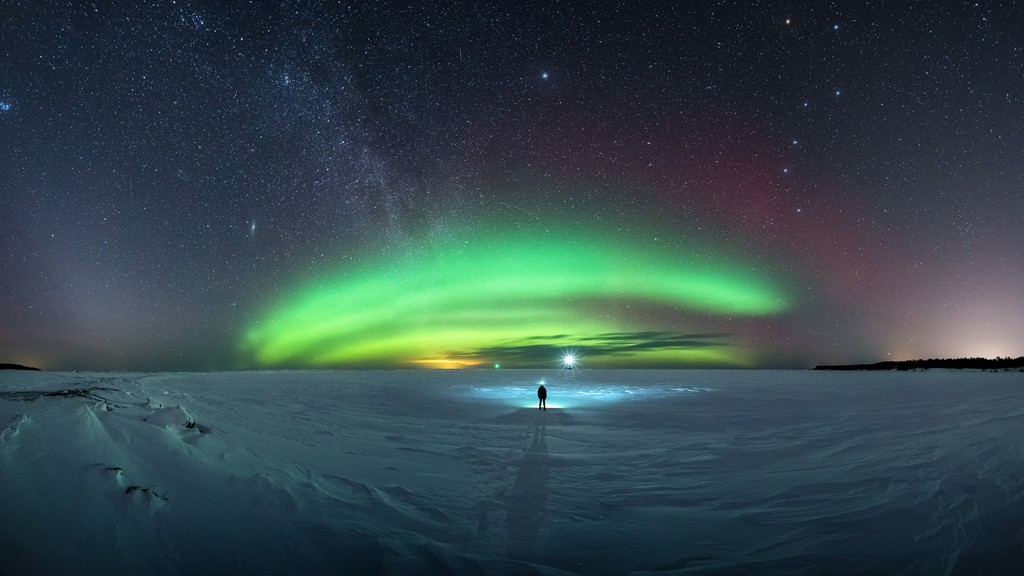 Apesar do frio, a chance de ver a brilhante Aurora Boreal atraiu esse observador do céu para a superfície congelada do Lago Superior, na costa oeste da Península de Keweenaw, nos Estados Unidos. Nesta composição panorâmica de imagens, feita em março de 2019, vê-se a distante Galáxia de Andrômeda (M31) e a faixa celeste da Via Láctea, à esquerda, brilhando acima do arco auroral esverdeado. A estrela próxima do Pólo Norte, Polaris - a principal da Constelação da Ursa Menor - está centrada acima e acompanhada, à direita, pelo asterismo da Constelação da Ursa Maior. As luzes terrestres incluem marcadores para dois quebra-mares no horizonte, perto do centro da cena