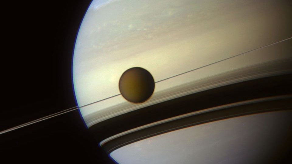 A espaçonave Cassini registrou esta imagem de Titã, a maior lua de Saturno, que parece ter um bronzeado indefinido, pois está continuamente envolta em nuvens espessas. Os anéis de Saturno são vistos como uma linha fina porque são muito planos e foram fotografados quase de lado. Os detalhes dos anéis de Saturno estão mais visíveis nas sombras escuras, vistas no topo das nuvens do planeta gigante. Como as partículas do anel orbitam no mesmo plano de Titã, elas parecem espetar a lua em primeiro plano. No hemisfério superior de Saturno, as nuvens apresentam quedas em longas faixas brilhantes, indicando perturbações numa corrente de jato de alta altitude. Medições recentes de Titã sugerem que vastos oceanos de água podem existir em suas profundezas