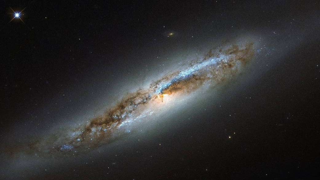 A Constelação de Virgem é especialmente rica em galáxias, devido especialmente à presença de uma coleção massiva e ligada gravitacionalmente de mais de 1,3 mil galáxias chamada Aglomerado de Virgem. Um membro específico desta comunidade cósmica, NGC 4388, aparece nesta imagem feita pelo Telescópio Espacial Hubble. Localizada a cerca de 60 milhões de anos-luz de distância, NGC 4388 está passando por uma transformação. Embora os arredores da galáxia pareçam suaves, como em galáxias elípticas, o seu centro exibe notáveis ​​faixas de poeira, em dois braços espirais simétricos , o que a torna uma galáxia espiral. Nesses braços, manchas azuis brilhantes marcam a localização de estrelas jovens