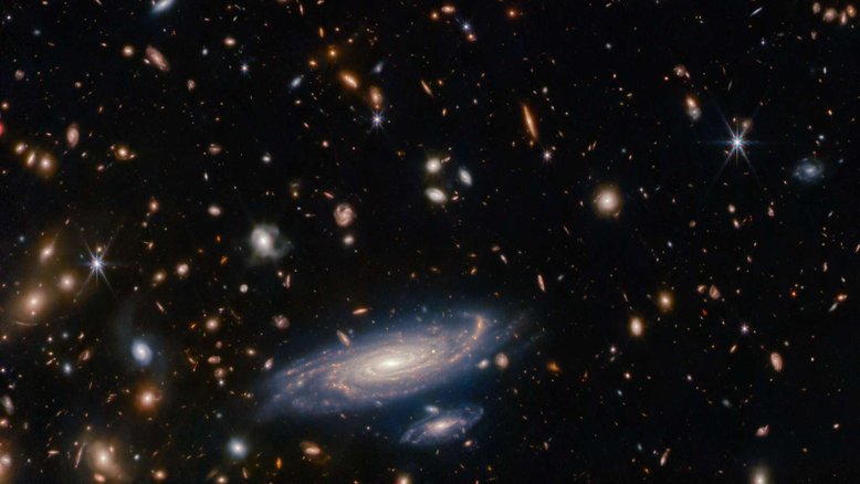 Um campo lotado de galáxias aglomera-se nesta imagem, registrada pelo Telescópio Espacial James Webb, junto a estrelas que brilham com os picos de difração característicos dos registros gerados por este moderno equipamento. A grande galáxia espiral, na parte inferior, foi batizada de LEDA 2046648 e está situada a pouco mais de um bilhão de anos-luz da Terra, na Constelação de Hércules. Um dos principais objetivos científicos do Webb é observar galáxias distantes — e, portanto, antigas — para compreender os detalhes da sua formação, evolução e composição