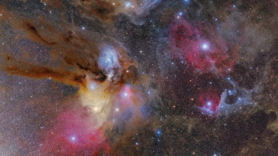 As nuvens coloridas que cercam o sistema estelar Rho Ophiuchi compõem uma das regiões de formação estelar mais próximas da Terra. Rho Ophiuchi é um sistema estelar binário, visível na nebulosa de reflexão azul, à esquerda do centro da imagem. Localizado a cerca de 400 anos-luz de distância, este sistema estelar distingue-se pelos seus arredores multicoloridos, que incluem uma nebulosa de emissão vermelha e numerosas faixas de poeira, nas cores castanho claro e escuro. Embora a maioria de suas características sejam visíveis por meio de um telescópio apontado para a região que compreende a Constelação do Serpentário, a Constelação de Escorpião e a Constelação de Sagitário, a única maneira de ver os detalhes intrincados dos redemoinhos de poeira, como na imagem acima, é com o auxílio de uma câmara de longa exposição