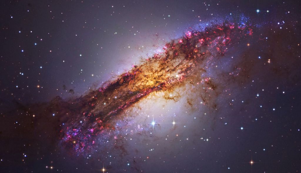 Qual é a galáxia ativa mais próxima do planeta Terra? É Centaurus A, a apenas 11 milhões de anos-luz de distância. Abrangendo mais de 60.000 anos-luz, a peculiar galáxia elíptica também é conhecida como NGC 5128. Formada a partir da colisão de duas galáxias, ela exibe diversos aglomerados de jovens estrelas azuis, regiões rosadas de formação de estrelas e imponentes faixas de poeira escura que são vistas nesta imagem composta com dados de telescópios espaciais e terrestres. Perto do centro da galáxia, os restos cósmicos que sobraram são constantemente consumidos por um buraco negro central com bilhões de vezes a massa do Sol