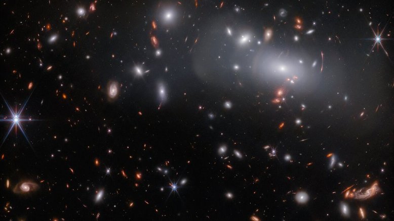 Esta imagem, feita pelo Telescópio Espacial James Webb, apresenta o enorme aglomerado de galáxias RX J2129. Devido às lentes gravitacionais, esta observação contém três imagens diferentes da mesma galáxia. As lentes gravitacionais ocorrem quando um corpo celeste é massivo o suficiente para curvar o espaço-tempo, dobrando a luz que passa por ele, como uma grande lente. Neste caso, a lente é o próprio aglomerado RX J2129, localizado a cerca de 3,2 bilhões de anos-luz da Terra, na Constelação de Aquário. As lentes gravitacionais podem fazer com que os objetos de fundo fiquem estranhamente distorcidos, como pode ser visto pelos arcos concêntricos de luz no canto superior direito desta imagem