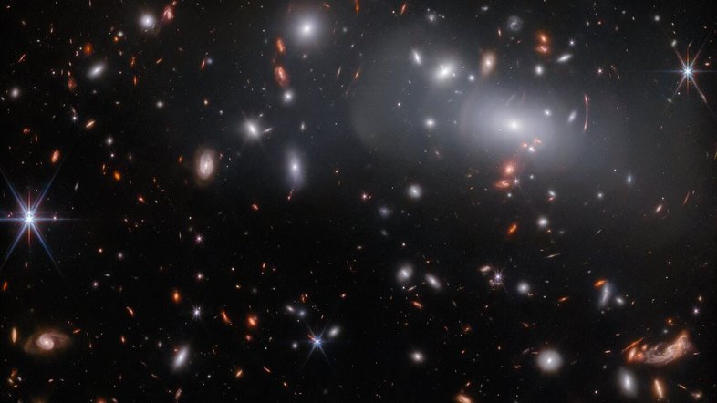Esta imagem, feita pelo Telescópio Espacial James Webb, apresenta o enorme aglomerado de galáxias RX J2129. Devido às lentes gravitacionais, esta observação contém três imagens diferentes da mesma galáxia. As lentes gravitacionais ocorrem quando um corpo celeste é massivo o suficiente para curvar o espaço-tempo, dobrando a luz que passa por ele, como uma grande lente. Neste caso, a lente é o próprio aglomerado RX J2129, localizado a cerca de 3,2 bilhões de anos-luz da Terra, na Constelação de Aquário. As lentes gravitacionais podem fazer com que os objetos de fundo fiquem estranhamente distorcidos, como pode ser visto pelos arcos concêntricos de luz no canto superior direito desta imagem