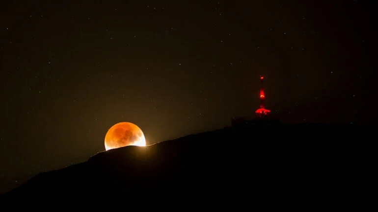 No dia 16 de julho de 2019, a celebração do 50º aniversário do lançamento da Missão Apollo 11, à Lua, contou com um Eclipse Lunar Parcial, que foi visível em diversos lugares da superfície terrestre. Nesta imagem, parte do disco lunar está imerso na sombra umbral escura e avermelhada da Terra. Esta paisagem noturna foi registrada após o nascer da Lua, na região Sul de Innsbruck, na Áustria, ao lado de uma torre de comunicação iluminada na cordilheira. Como os eclipses nunca ocorrem isolados, este Eclipse Lunar Parcial aconteceu na Lua Cheia após a Lua Nova, de 2 de julho do mesmo ano, na qual ocorreu também um Eclipse Solar Total