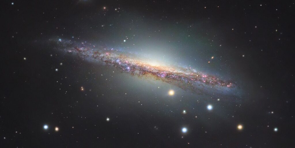 Grandiosa, a bela galáxia espiral NGC 1055 é o membro dominante de um pequeno grupo de galáxias a apenas 60 milhões de anos-luz de distância, na direção da Constelação de Cetus. Vista de lado, ela abrange mais de 100 mil anos-luz, sendo um pouco maior que a Via Láctea. As estrelas coloridas que aparecem na imagem estão em primeiro plano, dentro da nossa galáxia. As regiões rosadas em NGC 1055 demonstram a formação de estrelas. Com algumas galáxias de fundo ainda mais distantes, a imagem também revela um halo que se estende acima e abaixo do bulbo central de NGC 1055. O halo pode representar detritos de uma galáxia satélite perturbada pela espiral maior há cerca de 10 bilhões de anos
