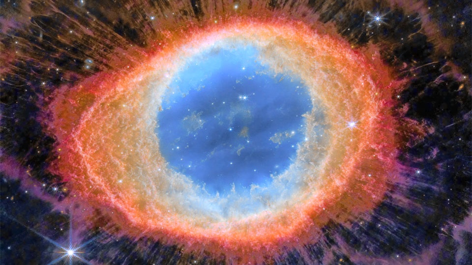 A Nebulosa do Anel (M57) é mais complicada do que parece quando vista de um pequeno telescópio. O anel central tem cerca de um ano-luz de diâmetro, mas esta notável exposição do Telescópio Espacial James Webb explora a nebulosa com luz infravermelha. Fios de gás, como cílios ao redor de um olho, tornam-se evidentes. Esses longos filamentos podem ser resultado do sombreamento de nós de gás denso em função da luz energética emitida em seu interior. A Nebulosa do Anel é uma nebulosa planetária alongada, um tipo de nuvem de gás criada quando uma estrela semelhante ao Sol evolui para se livrar de sua atmosfera externa e se tornar uma estrela anã branca. M57 fica a cerca de 2,5 mil anos-luz de distância, na Constelação da Lira