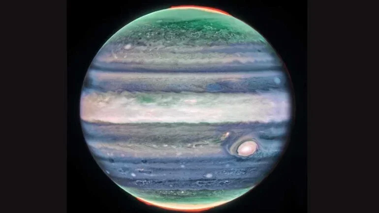 O Telescópio Espacial James Webb descobriu uma característica nova na atmosfera de Júpiter. Trata-se de uma corrente de jato de alta velocidade, que se estende por mais de 4,8 mil quilômetros de largura e fica sobre o equador do planeta, acima das principais plataformas de nuvens. Nesta imagem, em luz infravermelha, o brilho indica altitude elevada. As manchas e faixas brancas são provavelmente topos de nuvens de tempestades convectivas condensadas. As auroras, que aparecem em vermelho, estendem-se acima dos Pólos Norte e Sul do planeta