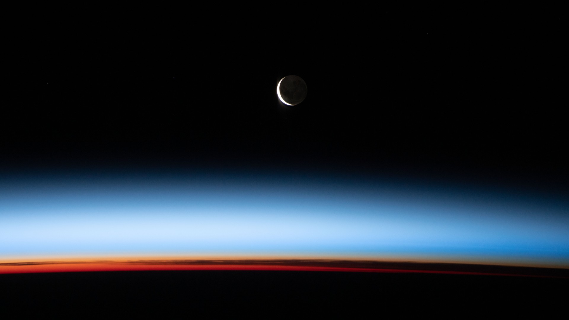 A Lua Nova está começando a crescer nesta imagem feita a partir da Estação Espacial Internacional (ISS). O registro aconteceu com o equipamento 431 quilômetros acima do Oceano Pacífico, a leste da Nova Zelândia. Vê-se uma fatia iluminada da Lua, enquanto a maior parte está voltada para o lado oposto ao Sol, ficando na escuridão. Acima da superfície da Terra, a sequência de cores retrata as várias camadas da atmosfera. Laranjas e amarelos profundos aparecem na troposfera, que contém mais de 80% da massa da atmosfera e quase todo o vapor d’água. A região rosa a branca, acima das nuvens, é a estratosfera inferior, com poucas ou nenhuma nuvem. Acima da estratosfera, as camadas azuis marcam a transição entre a atmosfera média e superior, gradualmente desaparecendo na escuridão do espaço sideral