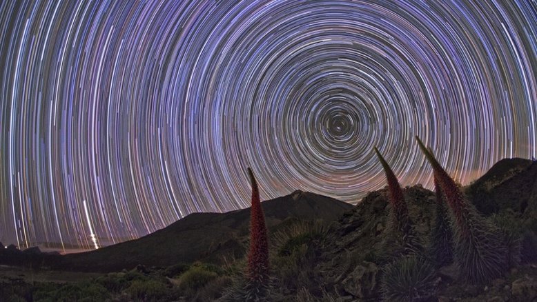 Nesta cena composta, o céu está repletoNesta cena composta, o céu está repleto de trilhas de estrelas ao redor do Polo Celeste Norte. Reflexo da rotação diária da Terra em seu eixo, os rastros de estrelas são familiares aos fotógrafos que fixam suas câmeras em um tripé e fazem longas exposições do céu noturno. As plantas de formas imponentes são tajinastes vermelhos, encontrados em Tenerife, nas Ilhas Canárias. Os tajinastes são plantas com flores raras que atingem uma altura de até 3 metros. Escondidos entre as rochas do terreno vulcânico, eles florescem na primavera e no início do verão, morrendo depois que suas sementes amadurecem. No horizonte distante, abaixo e à esquerda do polo celeste, vê-se o vulcão Teide de trilhas de estrelas ao redor do Pólo Celeste Norte. Reflexo da rotação diária da Terra em seu eixo, os rastros de estrelas são familiares aos fotógrafos que fixam suas câmeras em um tripé e fazem longas exposições do céu noturno. As plantas de formas imponentes são tajinastes vermelhos, encontrados em Tenerife, nas Ilhas Canárias. Os tajinastes são plantas com flores raras que atingem uma altura de até 3 metros. Escondidos entre as rochas do terreno vulcânico, eles florescem na primavera e no início do verão, morrendo depois que suas sementes amadurecem. No horizonte distante, abaixo e à esquerda do pólo celeste, vê-se o vulcão Teide