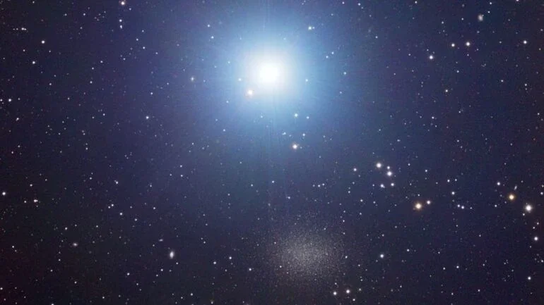 A estrela na parte superior é tão brilhante que às vezes é difícil notar a galáxia na parte inferior. Na imagem acima, pode-se ver a estrela Regulus e a galáxia Leão I, que ficam próximas uma da outra no céu, na direção da Constelação do Leão. Regulus compõe um sistema estelar múltiplo, com uma estrela dupla companheira, visível abaixo e à esquerda dessa jovem e azulada estrela da sequência principal. Leão I é uma galáxia anã esferoidal, membro do Grupo Local de galáxias, que é dominado pela nossa Via Láctea e a Galáxia de Andrômeda (M31). Acredita-se que Leão I seja a mais distante das várias pequenas galáxias satélites conhecidas que orbitam a nossa Via Láctea. Regulus (Alpha Leonis) está localizada a cerca de 75 anos-luz de distância, em contraste com Leão I, que está a aproximadamente 800 mil anos-luz de distância