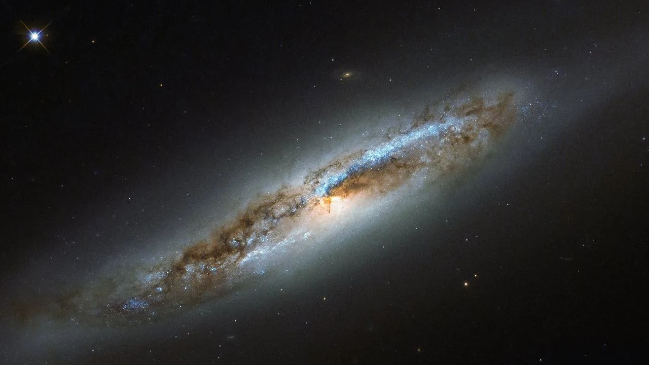 A Constelação de Virgem é muito rica em galáxias, especialmente devido à presença de uma coleção massiva e ligada gravitacionalmente de mais de 1,3 mil galáxias chamada Aglomerado de Virgem. Membro desse conjunto, NGC 4388 aparece nesta imagem do Telescópio Espacial Hubble. Localizada a cerca de 60 milhões de anos-luz de distância, NGC 4388 está experimentando transformações em função de interações gravitacionais com galáxias vizinhas, exibindo características de uma galáxia elíptica e também de uma galáxia espiral. Dentro dos braços, manchas azuis brilhantes marcam a localização de estrelas jovens, indicando que NGC 4388 hospedou recentes explosões de formação estelar