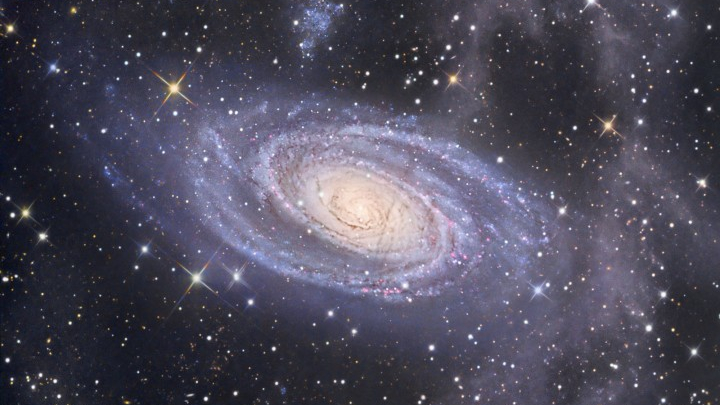 A galáxia espiral M81 fica a 11,8 milhões de anos-luz de distância, na Constelação da Ursa Maior. Esta imagem revela detalhes em seu núcleo amarelo brilhante, seus braços espirais azuis e extensas faixas de poeira. Acima de M81 encontra-se uma galáxia anã companheira, Holmberg IX, ostentando uma grande região rosada de formação de estrelas, perto do topo. Embora M81 e Holmberg IX estejam visíveis junto ao primeiro plano de estrelas na nossa galáxia, a Via Láctea, também é possível identificar nesta imagem um complexo mais tênue de nuvens de poeira. As nuvens relativamente inexploradas estão provavelmente a apenas algumas centenas de anos-luz de distância, bem acima do plano da nossa galáxia