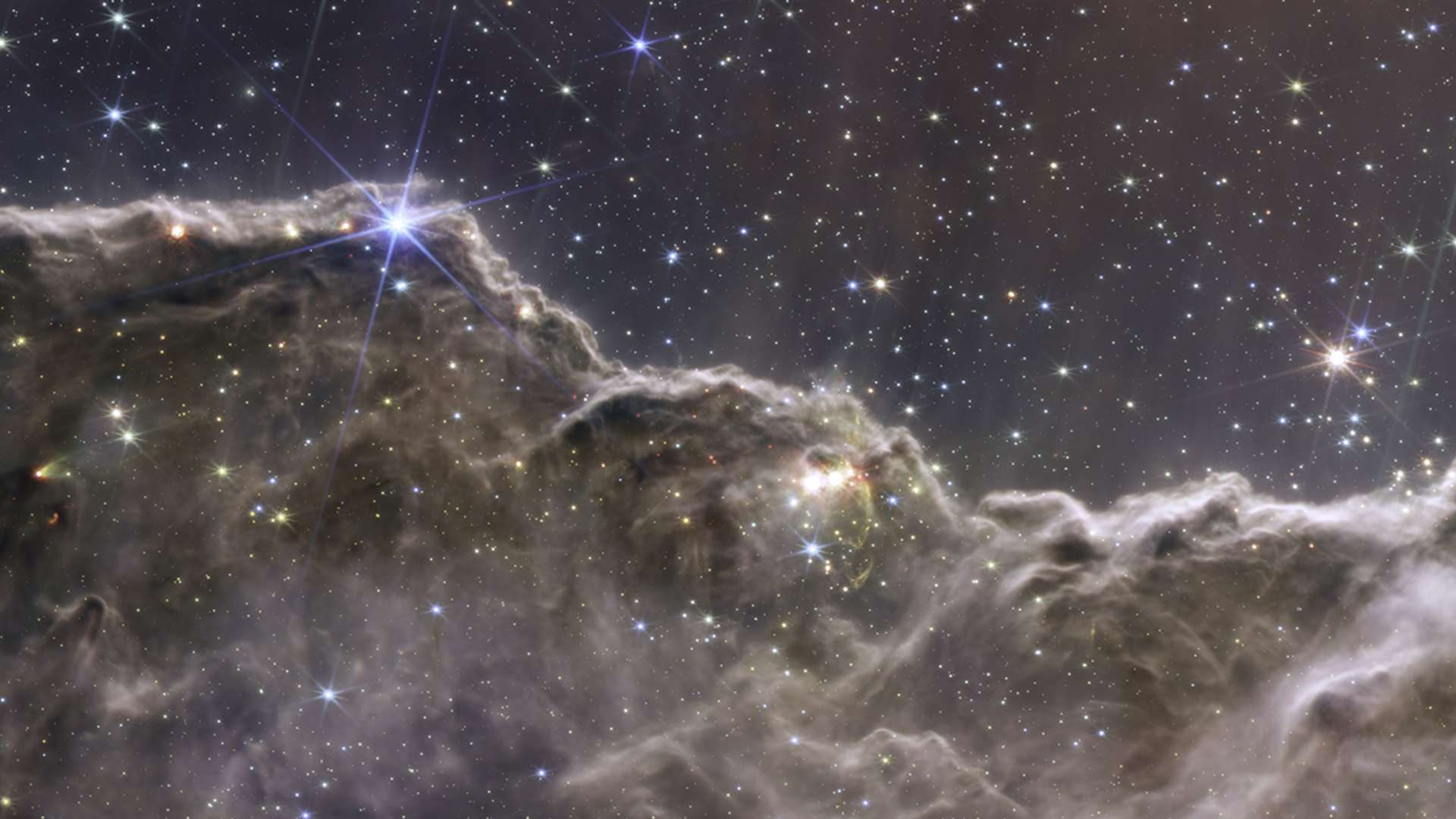 A combinação de imagens de diferentes câmeras do Telescópio Espacial James Webb permitiu criar esta imagem inédita de uma região de formação estelar na Nebulosa de Carina. Capturada em luz próxima ao infravermelho e infravermelho médio, este registro revela áreas anteriormente invisíveis do nascimento de estrelas. O que se parece com montanhas é, na verdade, a borda de uma região conhecida como NGC 3324. Esta gigantesca cavidade gasosa está a cerca de 7,6 mil anos-luz de distância, na direção da Constelação de Carina, e foi esculpida pela intensa radiação ultravioleta junto aos ventos estelares de estrelas jovens, extremamente massivas e quentes