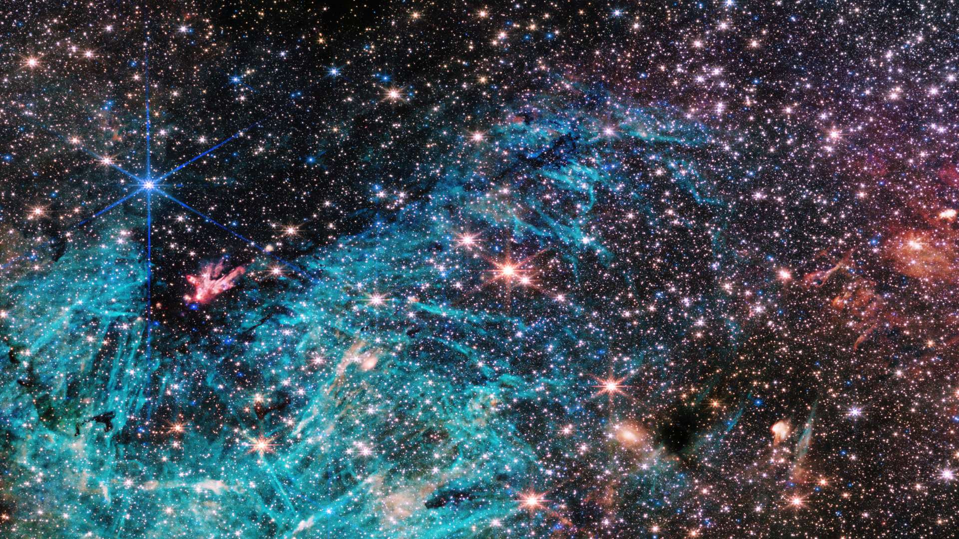 Esta visão do Telescópio Espacial James Webb revela uma porção de 50 anos-luz de largura do centro denso da Via Láctea, a nossa galáxia. Estima-se que 500 mil estrelas brilhem nesta imagem da região de Sagittarius C (Sgr C), a cerca de 300 anos-luz de distância do buraco negro supermassivo Sagittarius A*, que fica a aproximadamente 26 mil anos-luz de distância da Terra, na direção da Constelação de Sagitário. Uma vasta região de hidrogênio ionizado aparece em ciano, envolvendo uma nuvem infravermelha escura, que é tão densa a ponto de bloquear a luz de estrelas atrás dela